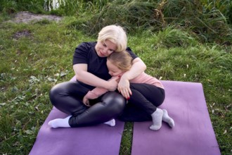 A mother and his daughter share a warm, comforting hug while sitting on yoga mats in a riverside.