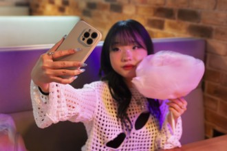 An Asian Gen Z girl captures a selfie with her phone while enjoying cotton candy in a vibrant