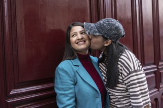 A loving Latin lesbian couple shares a sweet moment by a burgundy wall. One partner playfully