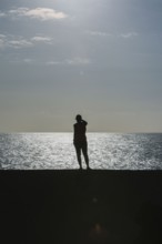 Silhouette of a person standing by the sea during sunset. The calm water and warm light create a