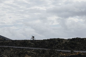 A lone cyclist riding on a volcanic road under a cloudy sky. The contrast between the dark terrain