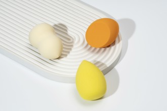 A trio of colorful cosmetic sponges in orange, yellow, and cream are arranged on a textured, white