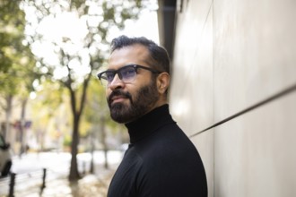 Side view of an Indian man dressed in a black turtleneck stands thoughtfully on a city street lined