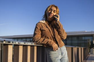 Mixed-race woman smiling while leaning on a railing and talking on a mobile phone outdoors She