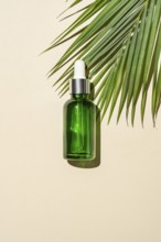 Top view of a green glass serum bottle with a dropper pipette, cast by a palm leaf's on a warm