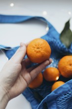 Cropped unrecognizable image of a hand holding a tangerine over a blue string bag filled with