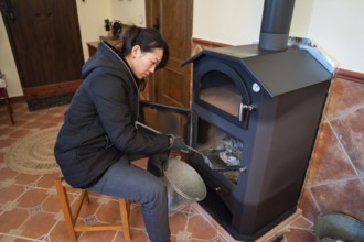 An Asian woman is shown sitting and cleaning a wood burning stove in a cozy indoor setting. She is