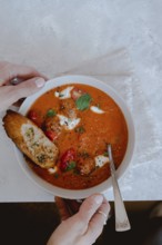 From above cropped unrecognizable person holding a bowl of tomato soup with meatballs, bread, and