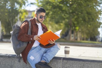 A latino transgender man reads a book in a sunny park He sits on a stone ledge with a backpack and