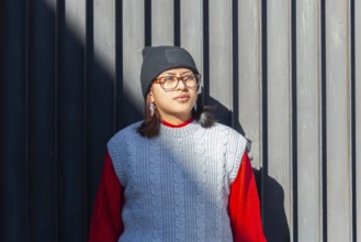 A Latina woman in stylish urban attire, including a knit vest and beanie, stands confidently by a