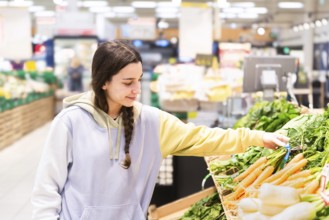 A young woman with a braid, wearing a casual hoodie, selects fresh vegetables in a brightly lighted