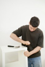 A young man in a casual t-shirt and jeans focuses intently on assembling a new white furniture