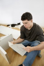 A young man sits on the floor attentively reading instructions for assembling a piece of white