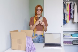 A young woman organizes clothes for online resale, using a laptop and smartphone. A box is ready