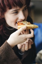 A young woman with red hair joyfully eats a slice of pizza, focusing on the flavor and her dining
