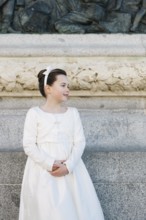 A young girl stands in a beautiful First Communion dress, framed by an ornate stone wall with