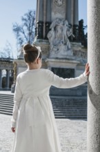 A young girl wearing a white First Communion dress stands beside a stone column, with a historic