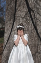 A young girl stands in front of a tree, covering her face with her hands. She is wearing an elegant