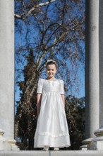 A young girl stands in a white First Communion dress, framed by stone columns and bare trees, with