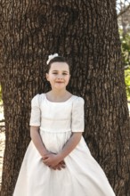 A young girl in a First Communion dress stands gracefully against a tree, embodying innocence and