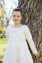 A young girl stands by a tree, wearing a white First Communion Day dress. She smiles softly under