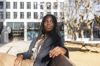 A young African American businesswoman with braids takes a relaxing break outdoors, sitting with a