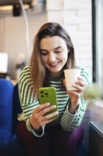A young woman in a striped green and white top happily operates her smartphone while enjoying a cup