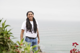 A man with long dreadlocks and tattoos smiles warmly, standing near vibrant flowers with the serene