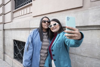 A Latin lesbian couple takes a joyful selfie outdoors, leaning against a historic building. Both