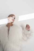 A confident middle-aged woman with a shaved head poses elegantly in a luxurious faux fur coat.