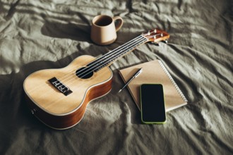 A warm morning scene featuring a ukulele, notebook, pen, smart phone, and coffee cup placed on a