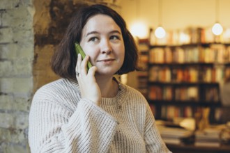 A young woman in a cream sweater smiles while talking on her green smartphone inside a warm, cozy