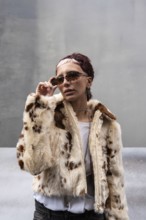 Stylish woman poses in a faux fur jacket and edgy eyewear. Urban setting enhances the chic look,