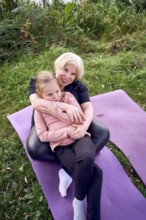 A joyful mother and daughter sit together on a yoga mat outdoors in a riverside. The lush, green
