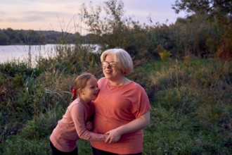A joyful mother and daughter share a laugh in a serene outdoor setting by the riverside at sunset.