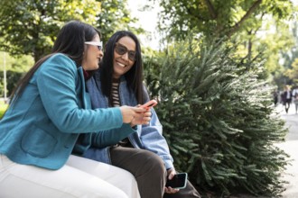 A joyful Latin lesbian couple sits on a park bench, sharing a moment with their smartphones under