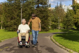 A young man walks alongside an elderly wheelchair user with cerebral palsy in a park, engaging in