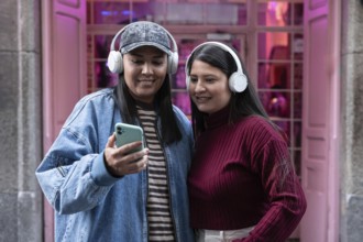 A latin lesbian couple listens to music with headphones while looking at a smartphone outdoors.