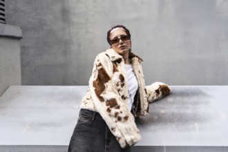Stylish woman wearing a cow print fur jacket, sunglasses, and denim, leaning against a metal