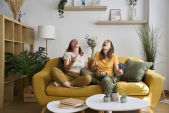 A joyful lesbian couple sits on a vibrant yellow sofa, tossing popcorn in the air and laughing