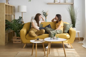 A cheerful lesbian couple enjoys a cozy moment on a yellow sofa The room is filled with greenery