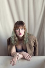 A woman with colorful hair leans on a sofa, wearing a brown blazer and eye-catching jewelry The