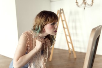 A woman in a lace top examines her reflection while wearing layered jewelry The minimalist room
