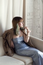 A stylish woman in a shimmering blue gown and brown coat sits thoughtfully on a beige couch Natural