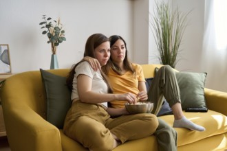 A lesbian couple relaxes on a yellow sofa, sharing popcorn and enjoying a quiet, intimate moment at