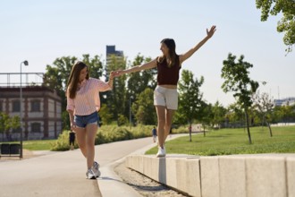 A joyful lesbian couple enjoys a sunny day, holding hands and balancing on a sidewalk in a park,