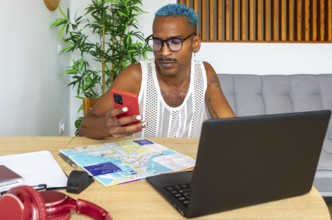A Hispanic LGBTQIA+ man plans a holiday. They're using a smartphone and laptop, surrounded by maps