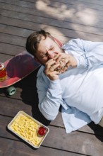A person enjoys a barbecue meal outdoors, lying beside a skateboard. The scene includes fries with
