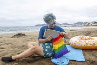 A transgender man with blue hair sitting on a beach, putting a tablet inside a rainbow pride bag.