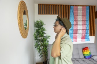 A young transgender person wearing a hat looking into a mirror reflection, stands near a couch in a